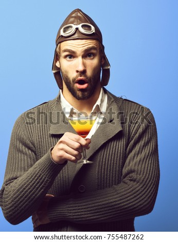 handsome bearded aviator man with long beard on surprised face holding glass of alcoholic cocktail in gray knitted sweater with hat and glasses on blue studio background