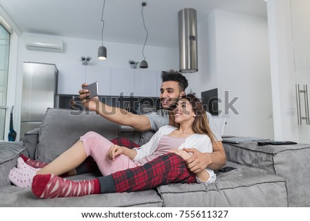 Happy Smiling Couple Taking Selfie Photo On Cell Smart Phones Sitting On Couch In Modern Apartment, Young Man And Woman Mke Self Portrait Embracing