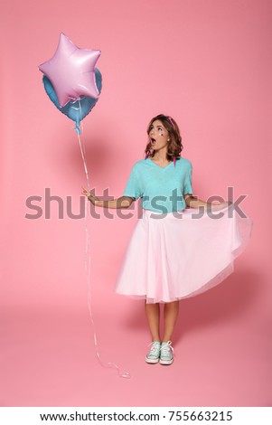 Full length portrait of an excited young girl in bright colorful clothes holding air balloons while standing and looking away isolated over pink background