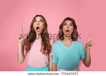 Close-up photo of two surprised woman in colorful tshirts pointing with finger and looking upward, isolated on pink background