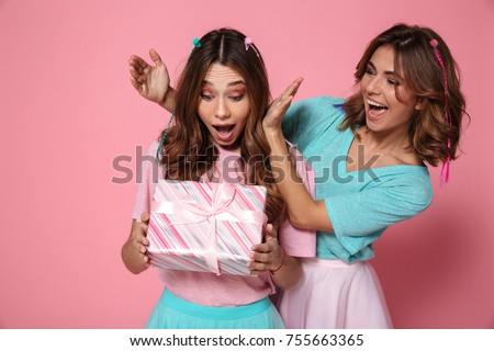 Young amazed woman holding present while celebrating birthday with her female friend, isolated over pink background