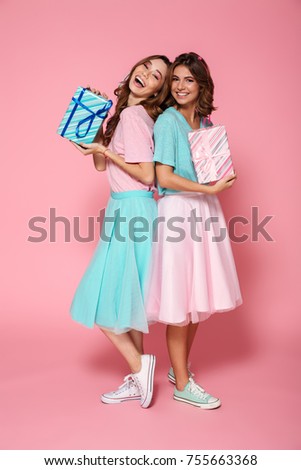 Two cheerful brunette woman in colorful clothes standing back to back and holding gift boxes, looking at camera, isolated on pink background
