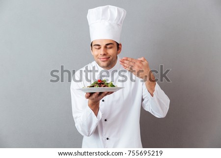 Picture of handsome young cook in uniform standing isolated over grey wall background holding salad and smell it.