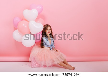 Happy brightful image of cute joyful little girl in tulle skirt sitting on present with balloons isolated on pink background. Amazing charming birthday fashionable kid looking to camera