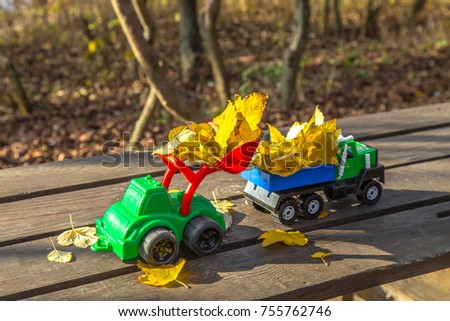 Two small toy truck and tractor is loaded with yellow fallen leaves. The car stands on a wooden surface against a background of a blurry autumn park. Cleaning and removal of fallen leaves. Seasonal wo