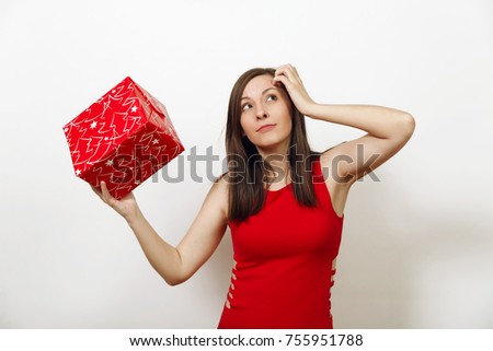 Interested engaged young woman dressed in red dress and Christmas hat holds gift box, thinks that she was given on white background. Santa girl with present isolated. New Year holiday 2018 concept