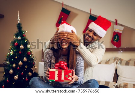 Happy playful smiling handsome young man sitting behind his girlfriend on the bed and holding her eye closed while she waiting to open Christmas gift.