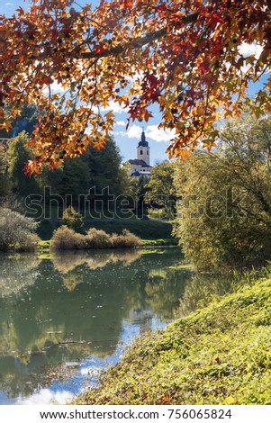 A beautiful view of St. Vid church in Ozalj Croatia through a red maple tree surrounded by forest, grass and lake taken on a sunny and cloudy autumn day