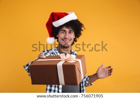 Portrait of a smiling cheerful african man dressed in christmas hat giving a gift box and looking at camera isolated over orange background