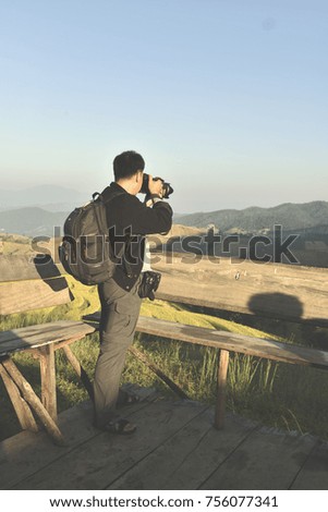 Happy Photographer Man in Winter Clothes With Camera Taking Landscape Photos on Old Wooden Terrace Top of Mountain Peak Against Beautiful Nature Green Mountain and Blue Sky.