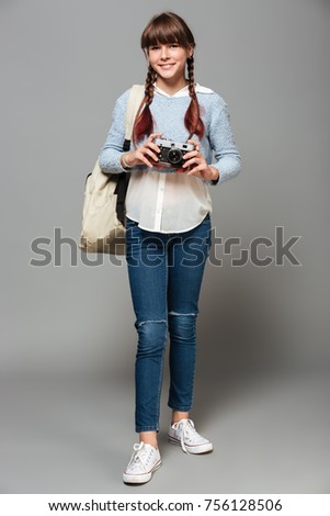 Full length portrait of a young cheerful schoolgirl with backpack holding photo camera and looking at camera isolated over gray background