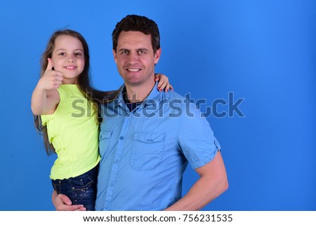 Girl and man with happy faces on blue background. Daughter and father hug each other. Schoolgirl sits on dads arms and shows thumbs up. Childhood and family concept.