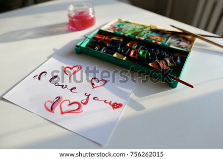 Lovely inscription I love you with picture of hearts in pink tones on white paper, written in calligraphic script that lies on light table with Colorful watercolor paints and glass jar with water