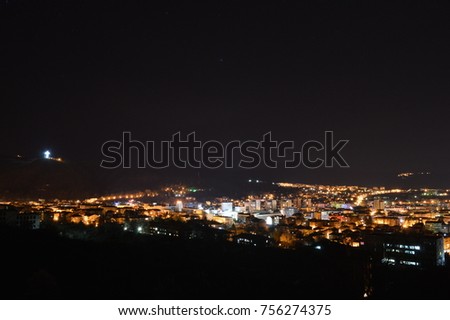 
man standing with his car on a hill overlooking a big city at night in the moonlight with the night lights of the city on a fog background in the sky and a stunning sensation
