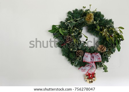 Home made Christmas wreath with holly, mistletoe, pine cones, ivy, tinsel, ribbon, artficial conifer twigs and bells against a light background with copy space to the left.