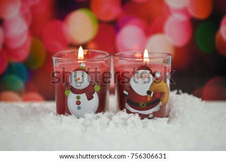 Warm cosy Christmas photography picture of glass red candles lit with yellow flame with Santa Claus and snowman design on laid in snow with pink fairy light background taken in England UK