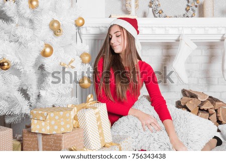 Young attractive brunette woman with gift box near Christmas tree and fireplace in cosiness home interior. Teenage girl in red santa hat. Winter holidays concept. White beautiful decor. Cute teenage .