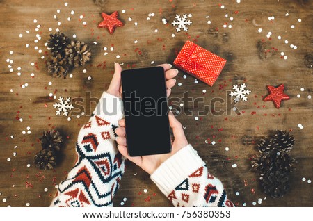 Female hands in a warm knitted sweater holding smart mobile phone with oled display on wooden background with Christmas gifts snowflakes and snow. Happy New Year and Xmas Flat lay composition top view Royalty-Free Stock Photo #756380353