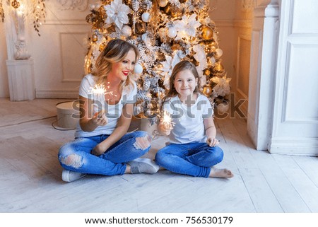Happy family mother and daughter with sparkler near a Christmas tree on Christmas eve at home. Woman and little girl relax in a white bedroom near the Christmas tree. Family at home