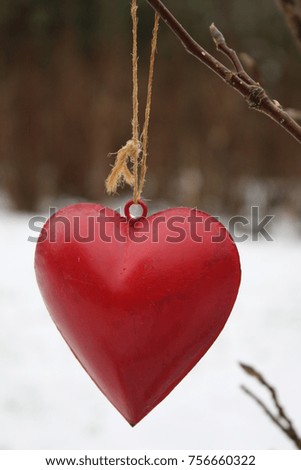 A red metal valentine love heart hanging from a branch on a garden tree with snow on the ground on a frosty spring day