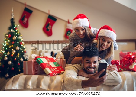 Three playful cute satisfied friends lying on the bed together and taking a selfie with Santa hats and gifts for Christmas holidays.