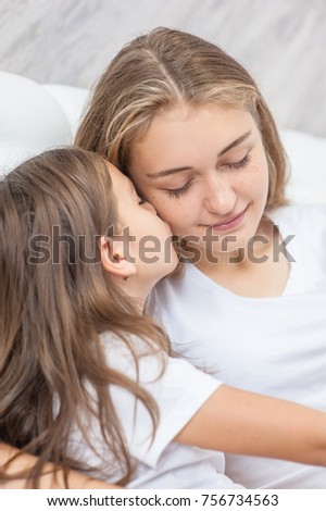  little girl is  kissing her beautiful young mom in cheek while sitting on bed at home