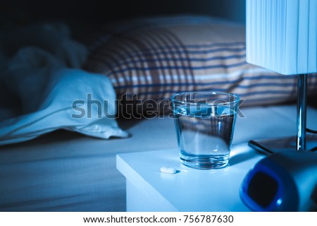 Sleeping pill on nightstand next to a glass of water. White tablet on table and bed in bedroom. Insomnia medicine concept. Royalty-Free Stock Photo #756787630