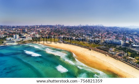 Wave breaks and surf on clear sand of famous Australian Sydney Bondi beach in aerial view with city CBD in background.