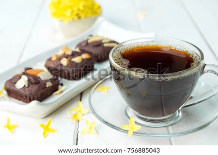 Hot coffee in clear cup and chocolate brownie cake in white plate on old white table , there is vase of beautiful yellow flower in the background