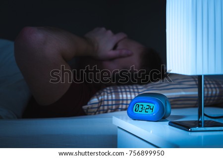 Insomnia and sleepless concept. Man unable to sleep. Exhausted and tired. Covering face with hand. Alarm clock on nightstand and bed in bedroom. Royalty-Free Stock Photo #756899950