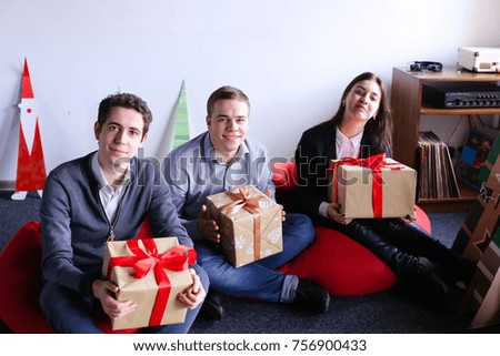 Happy friends in festive mood look away with smile and hold in their hands big Christmas boxes with gifts tied with red bows, sitting in soft red unusual armchairs near cardboard tree with garland and