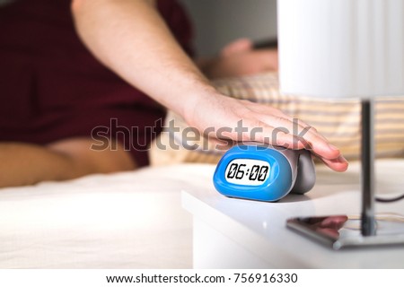 Man waking up in bed. Turning off alarm clock on nightstand or pressing snooze. Royalty-Free Stock Photo #756916330