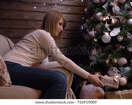 A beautiful blonde girl puts gifts under a Christmas tree. Beautiful jewelry and toys, light garland. 