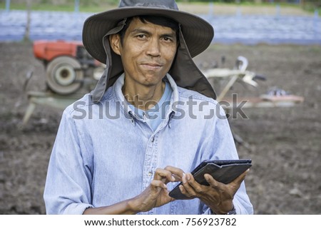 Asian farmer with hat using tablet in agriculture farm with cultivate machine in background