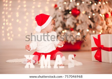 Cute baby girl wearing santa claus sitting on floor with white wooden xmas letters in room. Looking away. Holiday season. 