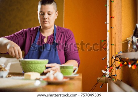 Beautiful young woman with short hair baking at home for christmas. She is sitting in the kitchen behind the table, mixing ingredients in the green bowl.