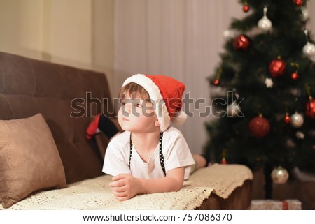Little boy with Santa hat sit on couch under a Christmas tree. Boy wait a New Year