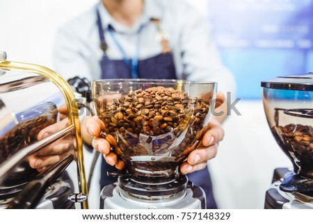 Barista man holding coffe grinder bowl with fresh roasted coffee beans in coffe shop or caffe bar. Selective focus, space for text.