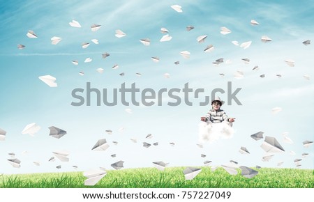 Young little boy keeping eyes closed and looking concentrated while meditating on cloud among flying paper planes with bright and beautiful landscape on background.