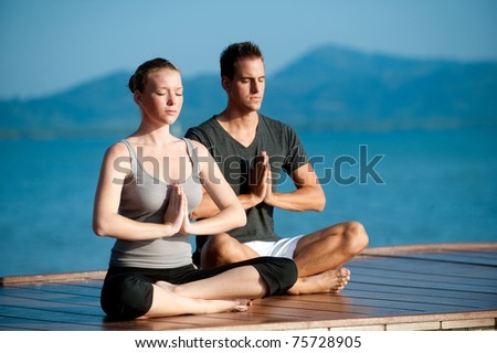 An attractive young woman and man doing yoga on a jetty with the blue ocean and another island behind them Royalty-Free Stock Photo #75728905