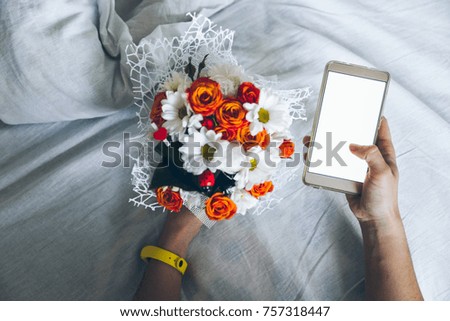 Hand holding mobile phone with flower bouquet white bed on background