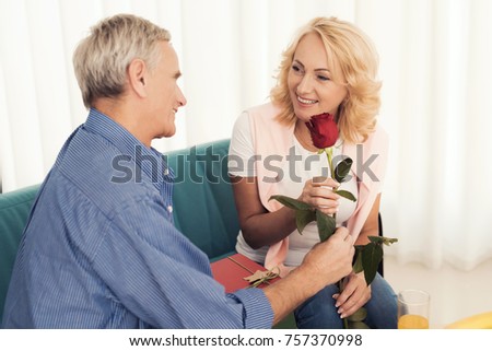 An elderly man gives a rose to an elderly woman. They are sitting on the couch. A woman is smiling at an elderly man. Next to the woman lies a gift.