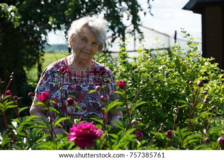 Portrait of old smiling woman in the park in the background of growing flowers as called peonies. Woman is charged energy of nature