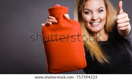 Positive smiling woman recommending warm hot water bottle in red soft fleece cover, on grey. Health care, pain relievers, recovery concept.