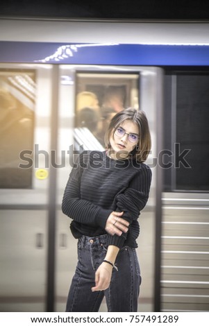 Teen female , girl with  glasses posing in subway railroad station .Fashion lady in sweater and denim jeans . Background of blurred extremely fast moving metro . Public transport style european.