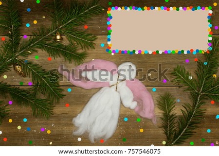 Christmas card with frame for text. One white woolen angel flying, colorful confetti and evergreen branches on wood plank.