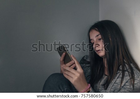  She is a victim of online  social networks. Sad teen checking phone sitting on the floor in the living room at home with a dark background. Victim of online bullying Stalker social networks