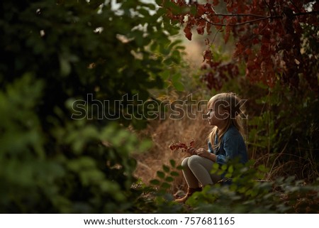 Little blond girl in bright dress and hat sits in a pine forest in sunny summer