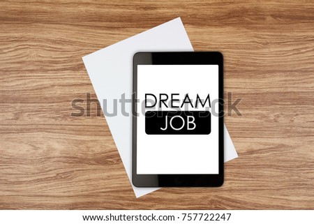 Digital tablet with text DREAM JOB on screen and white sheet on wooden desk