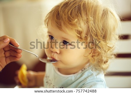 Cute fair-haired blond hazel-eyed kid little child baby boy fed with spoon eating photo portrait on blurred background, horizontal picture